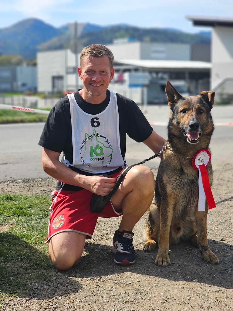 Hundeführer sitzt stolz neben seinem Deutschen Schäferhund und lächelt in die Kamera